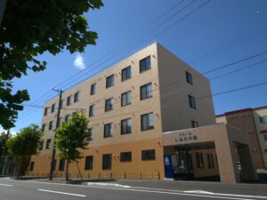 介護施設の建物外観。4階建ての淡いベージュ色の建築物で、1階に玄関スペースがある。